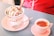 Two scoops of vanilla ice cream topped with chocolate syrup and almond slices are served in a colorful cup labeled with words like 'strawberry' and 'choco'. A wafer stick is placed in the ice cream. Next to it, there is a shot of espresso in a small pink cup on a matching saucer. A spoon lies beside the cup of ice cream on a pastel pink plate. The background features a red garment and a blue tabletop.
