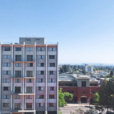 A multistory residential building with numerous windows stands prominently in an urban setting. Adjacent to it is another building with a red facade and a partial view of the cityscape in the background. Trees and greenery dot the landscape, and the sky is clear and bright blue.