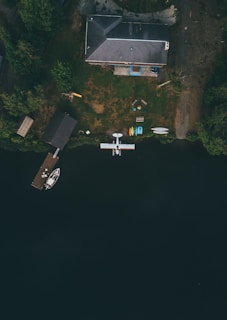 An aerial view captures a house with a grey roof situated near a calm body of water, surrounded by lush green trees. A dock extends into the water, with a small boat moored to it. Nearby, a seaplane floats, suggesting accessibility by air and water. The lawn in front of the house features various outdoor amenities, including a picnic table, chairs, and colorful kayaks.