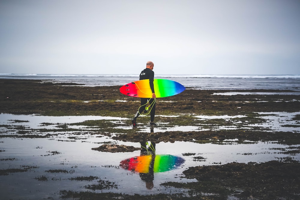 man in black wetsuit carrying red, yellow, blue, and green surfboard