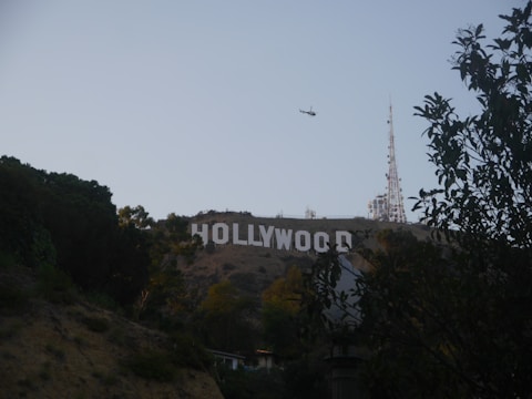 A hillside with the iconic Hollywood sign prominently displayed in large white letters. In the sky above, a helicopter hovers, and atop the hill, a tall communication tower is visible. Trees and shrubs fill the foreground, adding a touch of greenery, while the sky is clear and blue.
