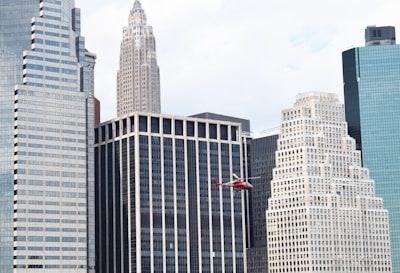 A red helicopter flies in front of a cluster of high-rise buildings in a city. The skyscrapers feature various architectural designs, including a mix of modern glass facades and more classic styles with ornate details. The sky is mostly clear, suggesting daytime.