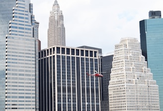 A red helicopter flies in front of a cluster of high-rise buildings in a city. The skyscrapers feature various architectural designs, including a mix of modern glass facades and more classic styles with ornate details. The sky is mostly clear, suggesting daytime.