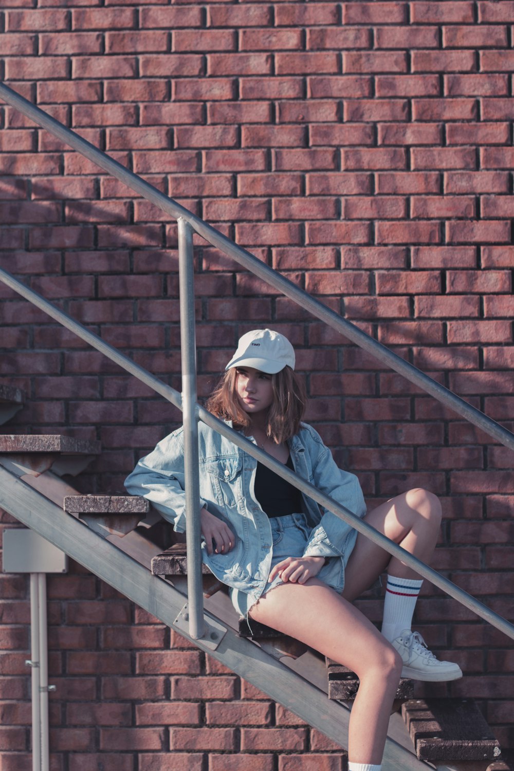 woman wearing gray denim jacket sitting on brown wooden stair near brown concrete wall during daytime