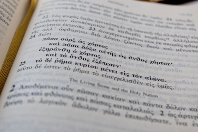 A close-up view of an open book with text in Greek. The page has an elegant ribbon bookmark in gold color visible on the left side. There are numbered sections of text, with larger numbers that appear to denote verses or chapters.