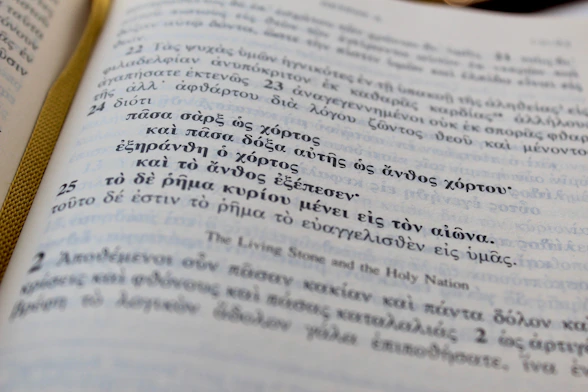 A close-up view of an open book with text in Greek. The page has an elegant ribbon bookmark in gold color visible on the left side. There are numbered sections of text, with larger numbers that appear to denote verses or chapters.