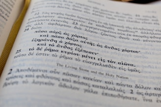A close-up view of an open book with text in Greek. The page has an elegant ribbon bookmark in gold color visible on the left side. There are numbered sections of text, with larger numbers that appear to denote verses or chapters.