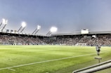 A large soccer stadium filled with spectators under bright floodlights. The field is green and well-maintained with players actively engaged in a match. Advertising banners line the perimeter of the stadium. A cameraman is positioned near the foreground, capturing the action on the field. The sky above is clear with a hint of dusk.