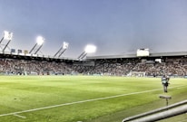 A large soccer stadium filled with spectators under bright floodlights. The field is green and well-maintained with players actively engaged in a match. Advertising banners line the perimeter of the stadium. A cameraman is positioned near the foreground, capturing the action on the field. The sky above is clear with a hint of dusk.