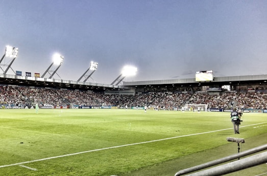 A large soccer stadium filled with spectators under bright floodlights. The field is green and well-maintained with players actively engaged in a match. Advertising banners line the perimeter of the stadium. A cameraman is positioned near the foreground, capturing the action on the field. The sky above is clear with a hint of dusk.