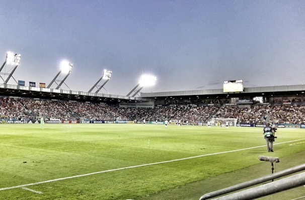A large soccer stadium filled with spectators under bright floodlights. The field is green and well-maintained with players actively engaged in a match. Advertising banners line the perimeter of the stadium. A cameraman is positioned near the foreground, capturing the action on the field. The sky above is clear with a hint of dusk.