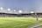A large soccer stadium filled with spectators under bright floodlights. The field is green and well-maintained with players actively engaged in a match. Advertising banners line the perimeter of the stadium. A cameraman is positioned near the foreground, capturing the action on the field. The sky above is clear with a hint of dusk.
