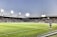 A large soccer stadium filled with spectators under bright floodlights. The field is green and well-maintained with players actively engaged in a match. Advertising banners line the perimeter of the stadium. A cameraman is positioned near the foreground, capturing the action on the field. The sky above is clear with a hint of dusk.