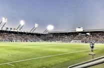 A large soccer stadium filled with spectators under bright floodlights. The field is green and well-maintained with players actively engaged in a match. Advertising banners line the perimeter of the stadium. A cameraman is positioned near the foreground, capturing the action on the field. The sky above is clear with a hint of dusk.