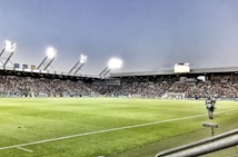 A large soccer stadium filled with spectators under bright floodlights. The field is green and well-maintained with players actively engaged in a match. Advertising banners line the perimeter of the stadium. A cameraman is positioned near the foreground, capturing the action on the field. The sky above is clear with a hint of dusk.