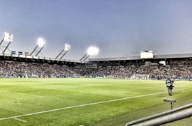 A large soccer stadium filled with spectators under bright floodlights. The field is green and well-maintained with players actively engaged in a match. Advertising banners line the perimeter of the stadium. A cameraman is positioned near the foreground, capturing the action on the field. The sky above is clear with a hint of dusk.