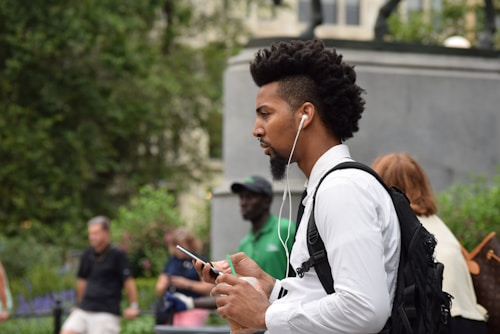 A man with a distinctive hairstyle listens to music through earphones while holding a smartphone. He wears a white shirt and carries a black backpack. The background includes blurred individuals and greenery.