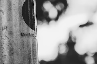 A black and white photograph featuring a close-up of a pole with the text 'Moreland City' partially visible on it. The background is blurred, creating a bokeh effect with indistinct shapes and light areas.