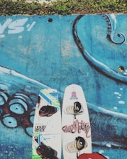 Two wakeboards with bindings are positioned on a blue surface with a detailed illustration that appears to be part of an octopus, showcasing its tentacles. There is green foliage along the top edge of the image.