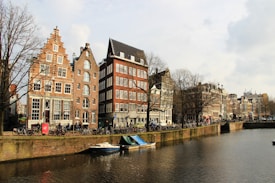 A picturesque canal lined with traditional Dutch gabled buildings, featuring a tranquil waterway with moored boats. Leafless trees line the street, bicycles are parked along the edge, and the day appears bright but chilly.