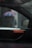 A car interior is viewed through a rainy window, with raindrops visible on the glass. Outside the car, a blurry figure is holding a multi-colored umbrella, likely providing shelter from the rain. The car's window and side mirror are prominently displayed.
