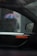 A car interior is viewed through a rainy window, with raindrops visible on the glass. Outside the car, a blurry figure is holding a multi-colored umbrella, likely providing shelter from the rain. The car's window and side mirror are prominently displayed.