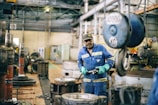 A worker wearing a blue uniform and green gloves is standing in an industrial workshop. The setting includes large machinery, metal parts, and a heavy-duty hook hanging from the ceiling. The space has a utilitarian and industrial atmosphere with concrete floors and a high ceiling.