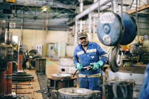 A worker wearing a blue uniform and green gloves is standing in an industrial workshop. The setting includes large machinery, metal parts, and a heavy-duty hook hanging from the ceiling. The space has a utilitarian and industrial atmosphere with concrete floors and a high ceiling.