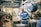 A worker wearing a blue uniform and green gloves is standing in an industrial workshop. The setting includes large machinery, metal parts, and a heavy-duty hook hanging from the ceiling. The space has a utilitarian and industrial atmosphere with concrete floors and a high ceiling.