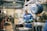 A worker wearing a blue uniform and green gloves is standing in an industrial workshop. The setting includes large machinery, metal parts, and a heavy-duty hook hanging from the ceiling. The space has a utilitarian and industrial atmosphere with concrete floors and a high ceiling.