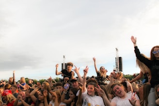 A lively crowd of people is gathered outdoors, enjoying a concert or music festival. Many individuals have their hands raised, some showing peace signs, and a few have their faces painted. There are large speakers in the background and a clear sky above.