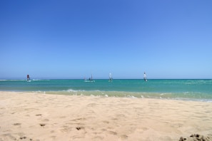 A picturesque beach scene with a wide expanse of sandy shore in the foreground leading to a calm turquoise sea. Several windsurfers are visible on the water, each with brightly colored sails. The sky above is clear and blue, creating a sense of openness and tranquility.
