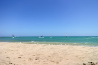 A picturesque beach scene with a wide expanse of sandy shore in the foreground leading to a calm turquoise sea. Several windsurfers are visible on the water, each with brightly colored sails. The sky above is clear and blue, creating a sense of openness and tranquility.
