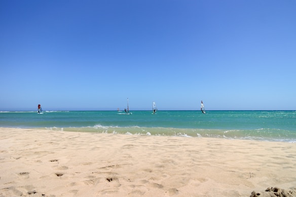 A picturesque beach scene with a wide expanse of sandy shore in the foreground leading to a calm turquoise sea. Several windsurfers are visible on the water, each with brightly colored sails. The sky above is clear and blue, creating a sense of openness and tranquility.