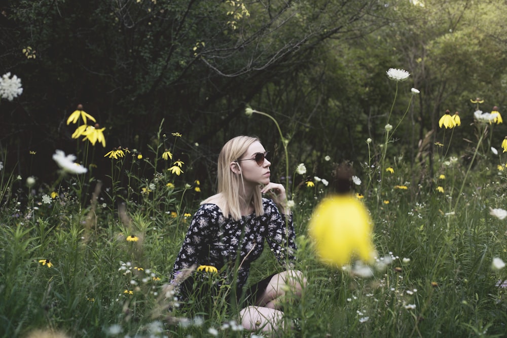 Mujer sentada en el campo de hierba rodeada de flores mientras la mano en la barbilla