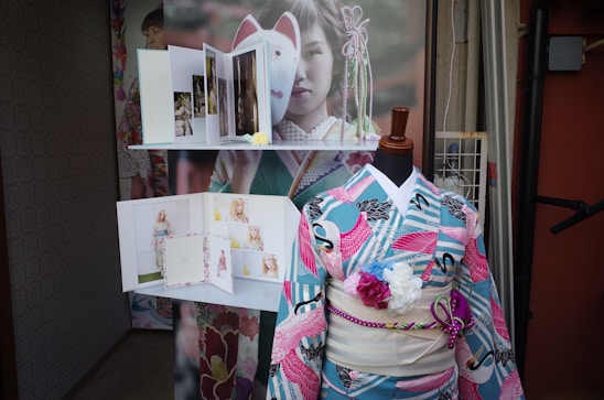A mannequin is dressed in a colorful kimono with a floral and crane pattern. Several photo albums are displayed on a shelf above, showcasing images of people in similar attire. A large poster behind the mannequin features a person in traditional dress.