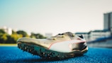 A track and field sports shoe is positioned on a blue running track, with soft sunlight illuminating its design. The shoe features sharp cleats underneath, a sleek metallic finish, and a signature inscribed on the side. Blurred buildings and greenery are visible in the background.