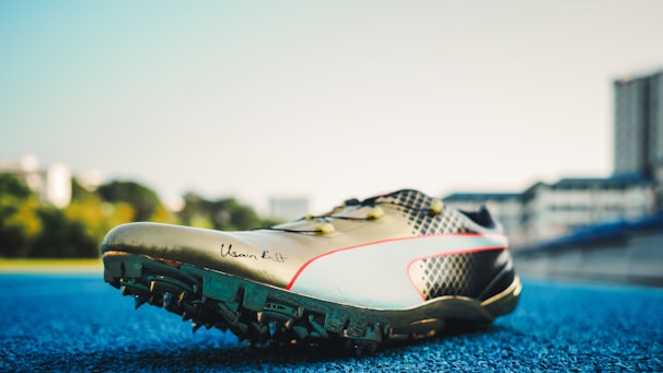 A track and field sports shoe is positioned on a blue running track, with soft sunlight illuminating its design. The shoe features sharp cleats underneath, a sleek metallic finish, and a signature inscribed on the side. Blurred buildings and greenery are visible in the background.