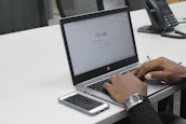 A person is using a laptop with a Google search page open on the screen. The laptop is placed on a white office desk, alongside a smartphone and a telephone. The person is typing on the keyboard and wearing a watch on their left wrist. The setting appears to be an office environment.