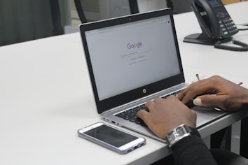 A person is using a laptop with a Google search page open on the screen. The laptop is placed on a white office desk, alongside a smartphone and a telephone. The person is typing on the keyboard and wearing a watch on their left wrist. The setting appears to be an office environment.