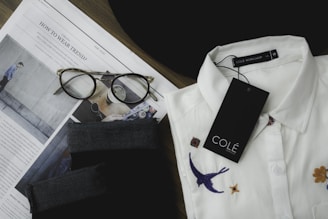 A neatly folded white collared shirt with embroidered designs, including a bird and flowers, is placed next to a pair of tortoiseshell eyeglasses on top of an opened fashion magazine. There is a black fabric item and a tag labeled 'COLÉ' on the shirt.
