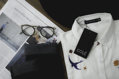 A neatly folded white collared shirt with embroidered designs, including a bird and flowers, is placed next to a pair of tortoiseshell eyeglasses on top of an opened fashion magazine. There is a black fabric item and a tag labeled 'COLÉ' on the shirt.