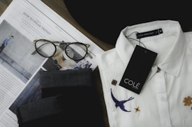 A neatly folded white collared shirt with embroidered designs, including a bird and flowers, is placed next to a pair of tortoiseshell eyeglasses on top of an opened fashion magazine. There is a black fabric item and a tag labeled 'COLÉ' on the shirt.