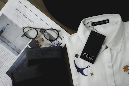 A neatly folded white collared shirt with embroidered designs, including a bird and flowers, is placed next to a pair of tortoiseshell eyeglasses on top of an opened fashion magazine. There is a black fabric item and a tag labeled 'COLÉ' on the shirt.