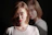 A person with a neutral expression is facing forward while their reflection appears in the background. They are wearing a white shirt and have light brown hair. The image has a dramatic, shadowed lighting effect.