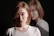 A person with a neutral expression is facing forward while their reflection appears in the background. They are wearing a white shirt and have light brown hair. The image has a dramatic, shadowed lighting effect.