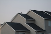 Several suburban houses with gabled roofs are aligned in a row under a gray sky. The houses are uniformly built with beige siding and black asphalt shingle roofs, giving a uniform and orderly appearance.