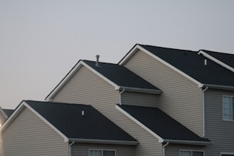 Several suburban houses with gabled roofs are aligned in a row under a gray sky. The houses are uniformly built with beige siding and black asphalt shingle roofs, giving a uniform and orderly appearance.