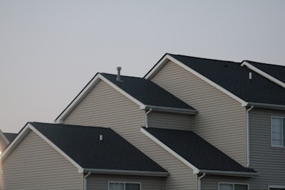 Several suburban houses with gabled roofs are aligned in a row under a gray sky. The houses are uniformly built with beige siding and black asphalt shingle roofs, giving a uniform and orderly appearance.