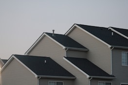 Several suburban houses with gabled roofs are aligned in a row under a gray sky. The houses are uniformly built with beige siding and black asphalt shingle roofs, giving a uniform and orderly appearance.